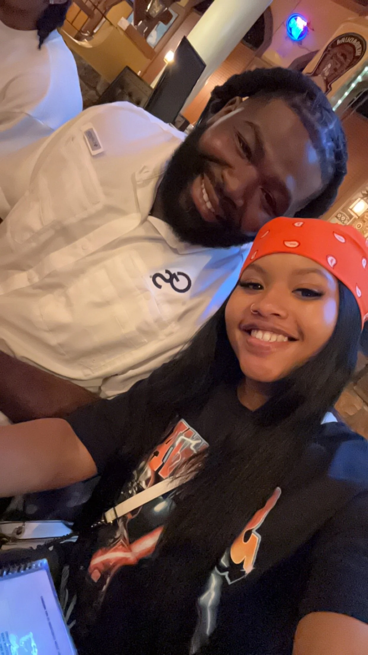 A man with a beard and long braided hair smiling, and a woman with long dark hair, wearing a red bandana and a black T-shirt with colorful graphics, taking a selfie together at a restaurant or bar with warm lighting and decorative signs in the background.