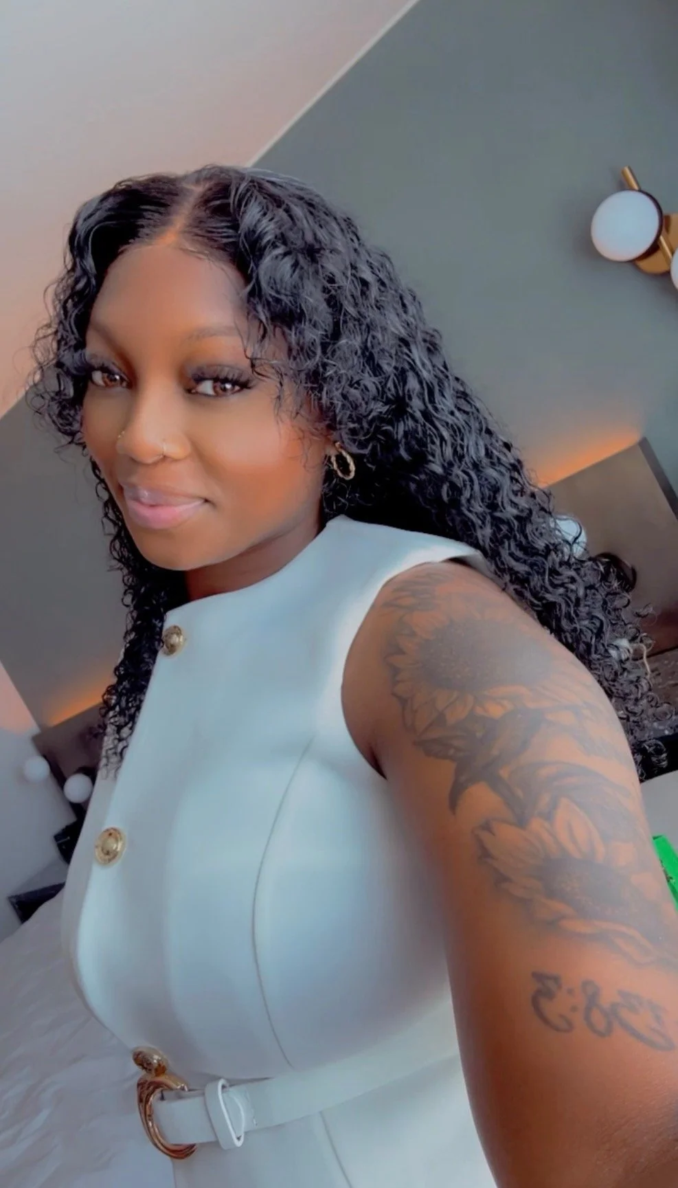 A woman with curly black hair taking a selfie in a hotel room, wearing a white sleeveless dress with gold buttons and a white belt, showing a floral tattoo on her upper arm.