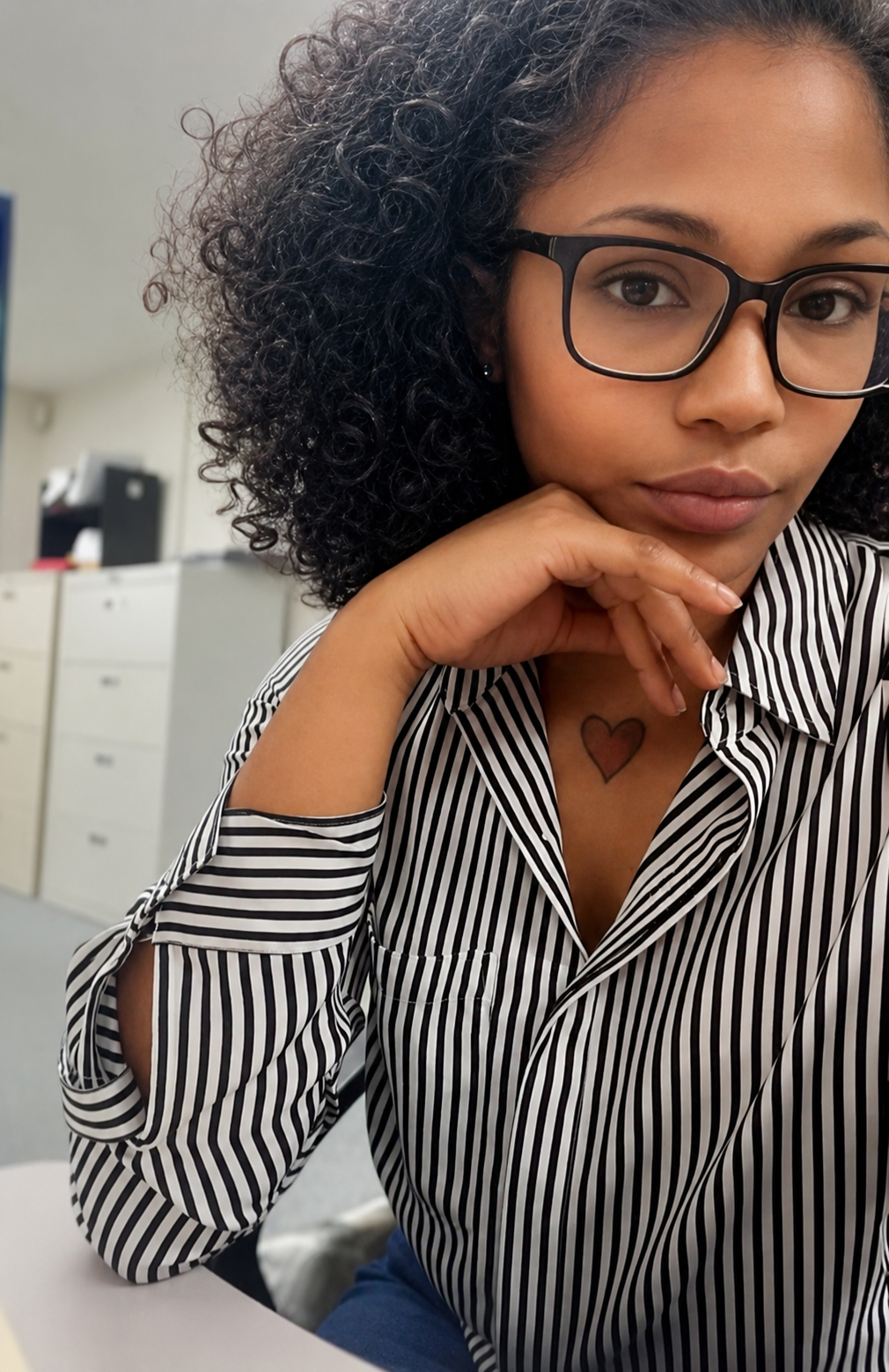 A young woman with curly hair, glasses, and a heart tattoo on her chest, wearing a black and white striped shirt, sitting in an office environment with her hand under her chin.