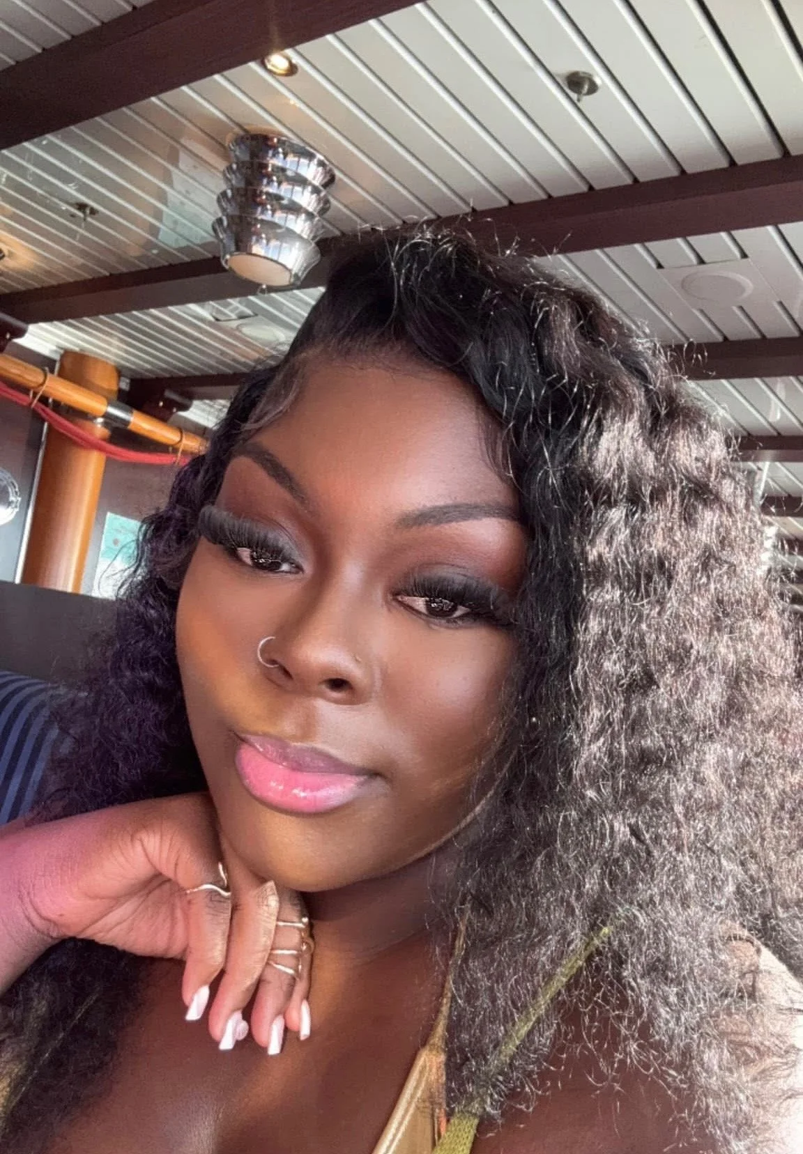 A woman with curly hair, makeup, and a nose ring, sitting at a table in a restaurant or cafe with a metallic decorative element on the ceiling.
