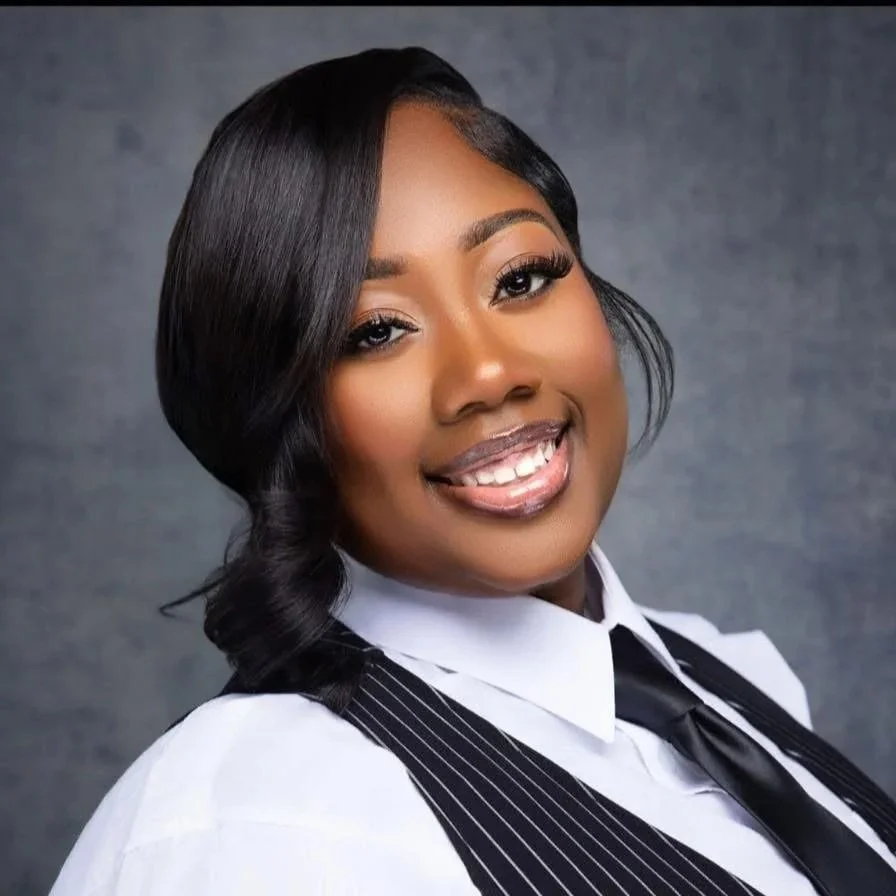 A young Black woman with shoulder-length black hair styled in loose waves, smiling and wearing a white collared shirt, a black-vin striped vest, and a black tie, against a gray background.