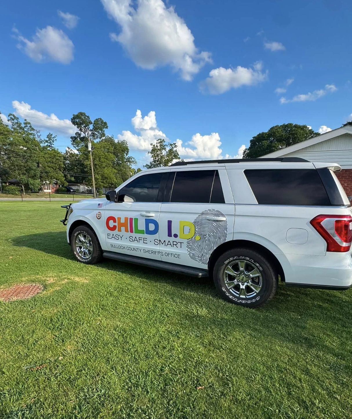 A white SUV parked on a grassy area under a blue sky with scattered clouds. The SUV has colorful text on the side that reads, 'CHILD I.D. EASY SAFE SMART,' and is associated with the Bulloch County Sheriff's Office.