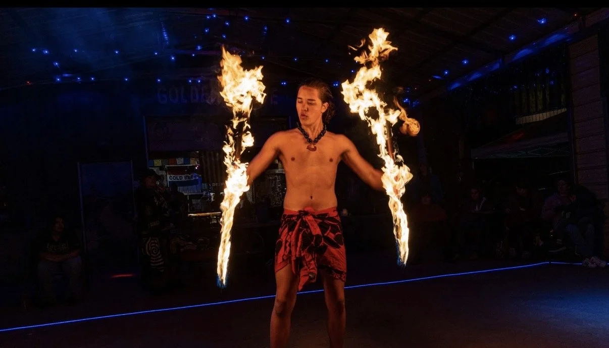 A Kaneli with a red lavalava and a necklace, spinning fire Knife Doubles at the Embers flow night with audience in the background.