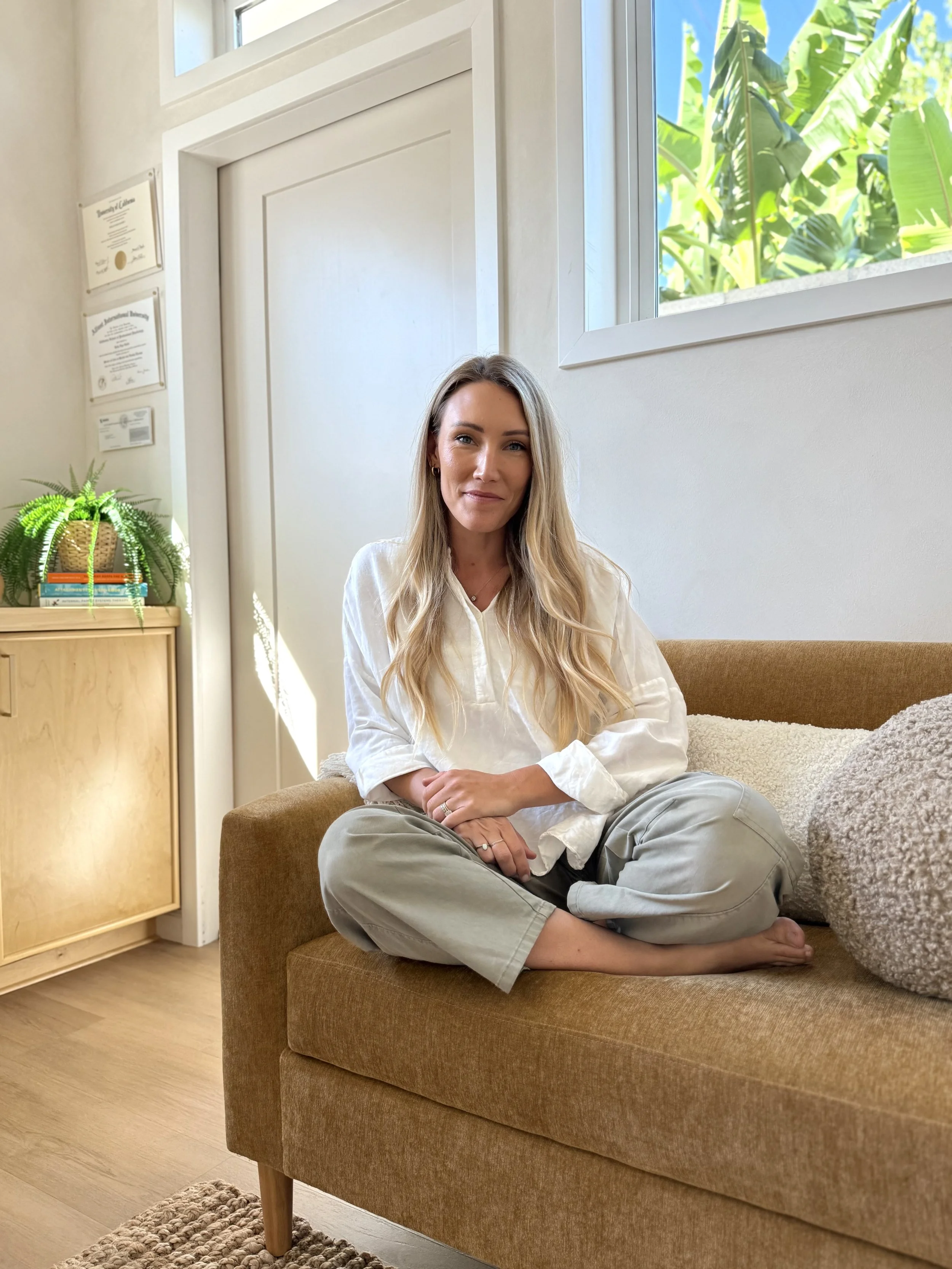 Therapist sitting cross-legged on a tan couch in a brightly lit therapy office with white walls, a window with greenery outside, a wooden cabinet with a potted fern and books, and framed certificates on the wall.