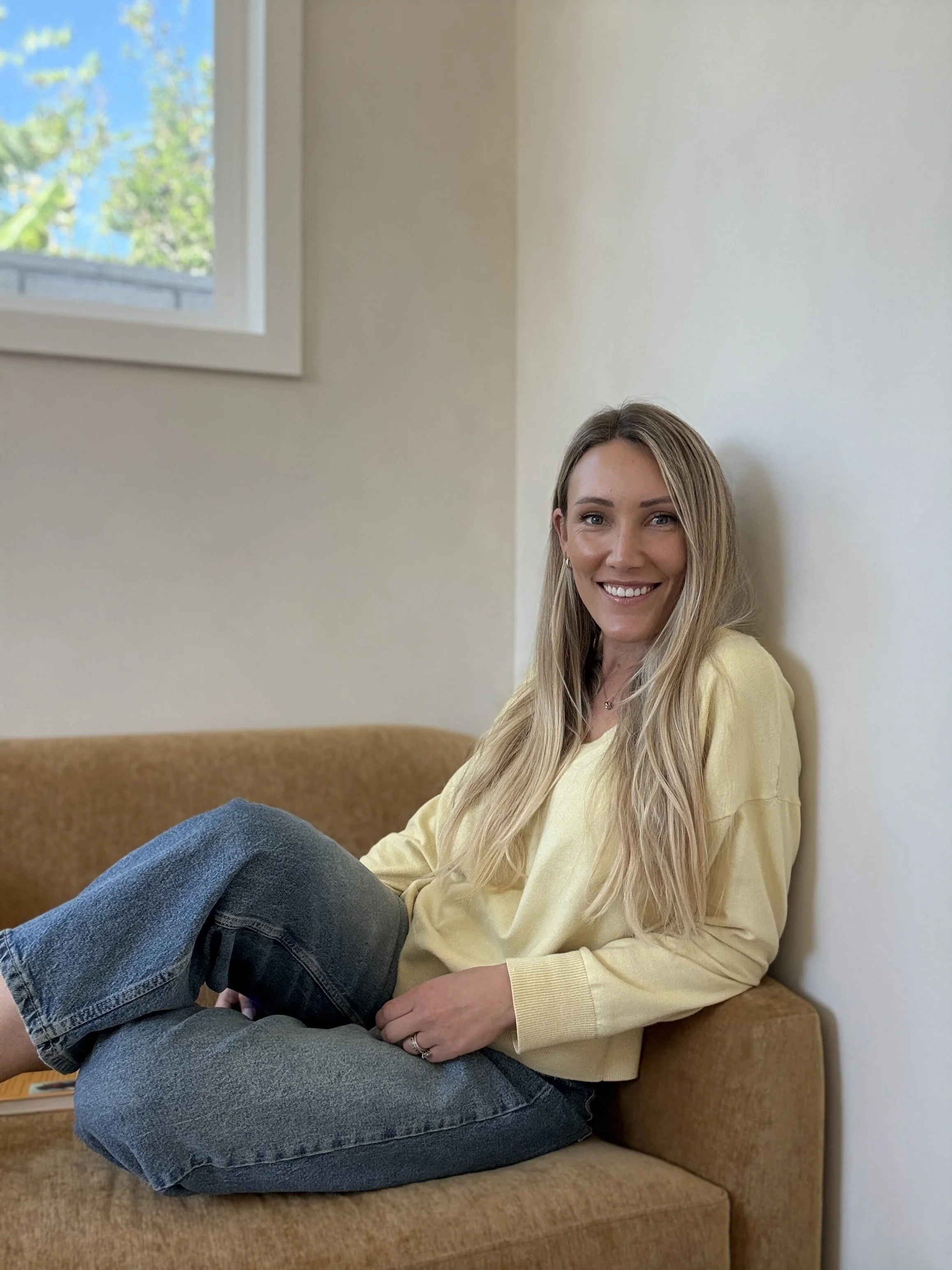 Welcoming therapist smiling, sitting on an orange sofa wearing a yellow sweatshirt and blue jeans in a brightly lit room.