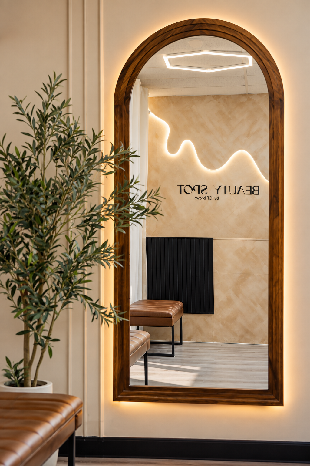 A large, arched wooden framed mirror reflecting a beige wall with a glowing abstract line art and the text 'BEAUTY SPOT by CT BROWS'. There are brown leather benches and a potted plant visible in the room.