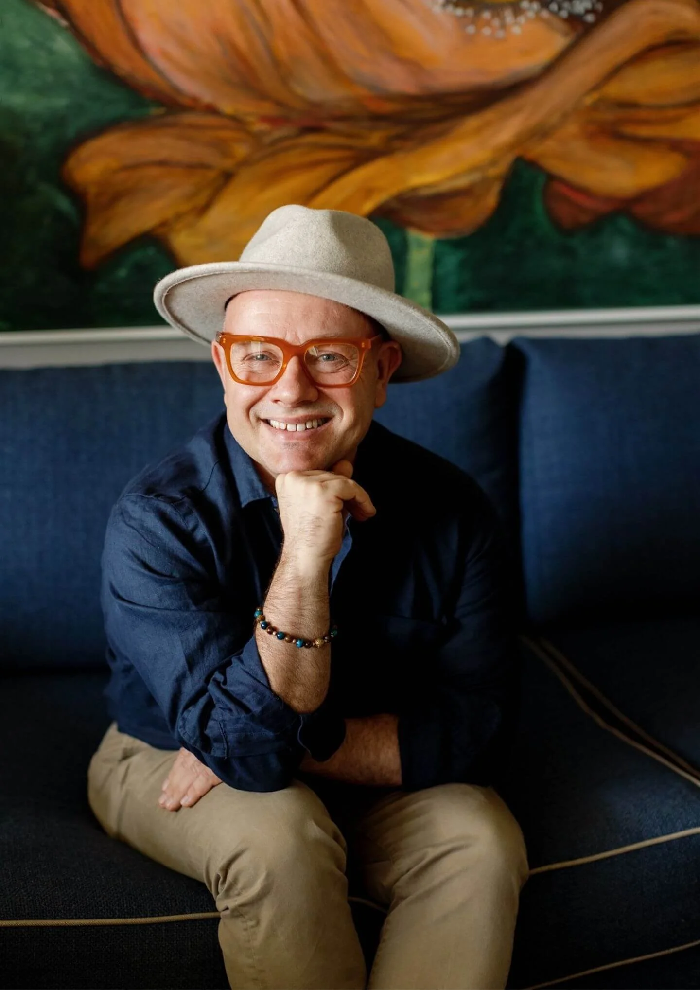 A smiling man wearing a beige hat, orange glasses, a dark blue shirt, and a beaded bracelet, sitting on a blue couch with a colorful painting of a pumpkin in the background.