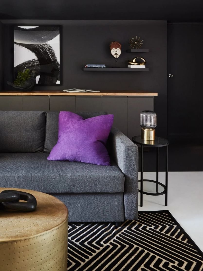 Contemporary living room with a gray sofa, purple pillow, black and white geometric rug, black side table with a decorative lamp, black wall with shelving, and artwork.
