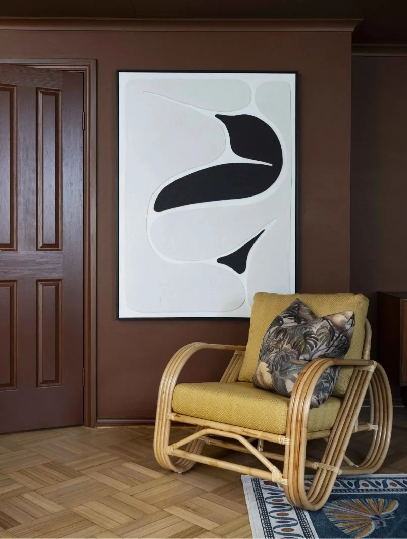 A room featuring a wooden door, a framed abstract black and white artwork on a brown wall, and a yellow armchair with a floral pillow.