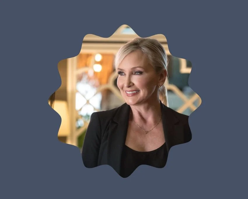 A smiling blonde woman in a black blazer and top, looking to her right, inside a warmly lit room.