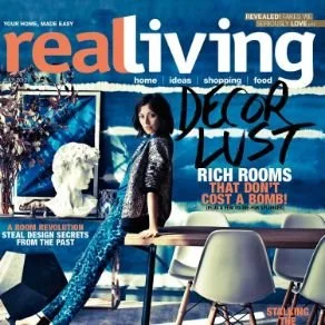 Cover of Real Living magazine featuring a woman standing in a modern dining room with a Christmas tree, a bust sculpture, and stylish furniture, with headlines about decor, rich rooms, and home secrets.