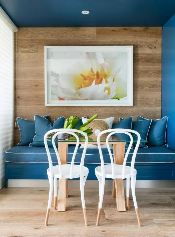 A cozy room with a blue cushioned bench with multiple matching pillows, a wall art of a white flower behind the bench, a wooden table with a flower arrangement, and two white chairs in front of the table. The room has blue walls and wooden flooring.