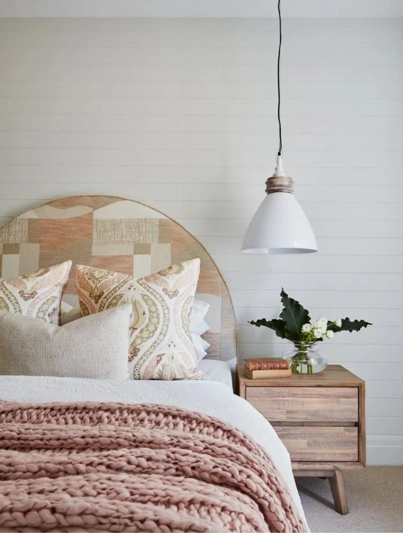 Minimalist bedroom with a wooden bed and pale pink chunky knit blanket, decorative pillows, a wooden nightstand with a glass vase of white flowers and an old book, white shiplap wall, and a white pendant light hanging from the ceiling.