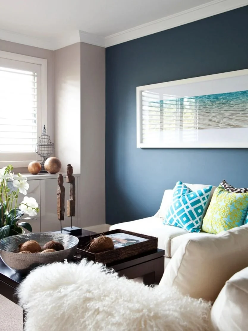 Living room with blue accent wall, white sofa with colorful patterned pillows, coffee table with decorative items, and a window with white blinds.
