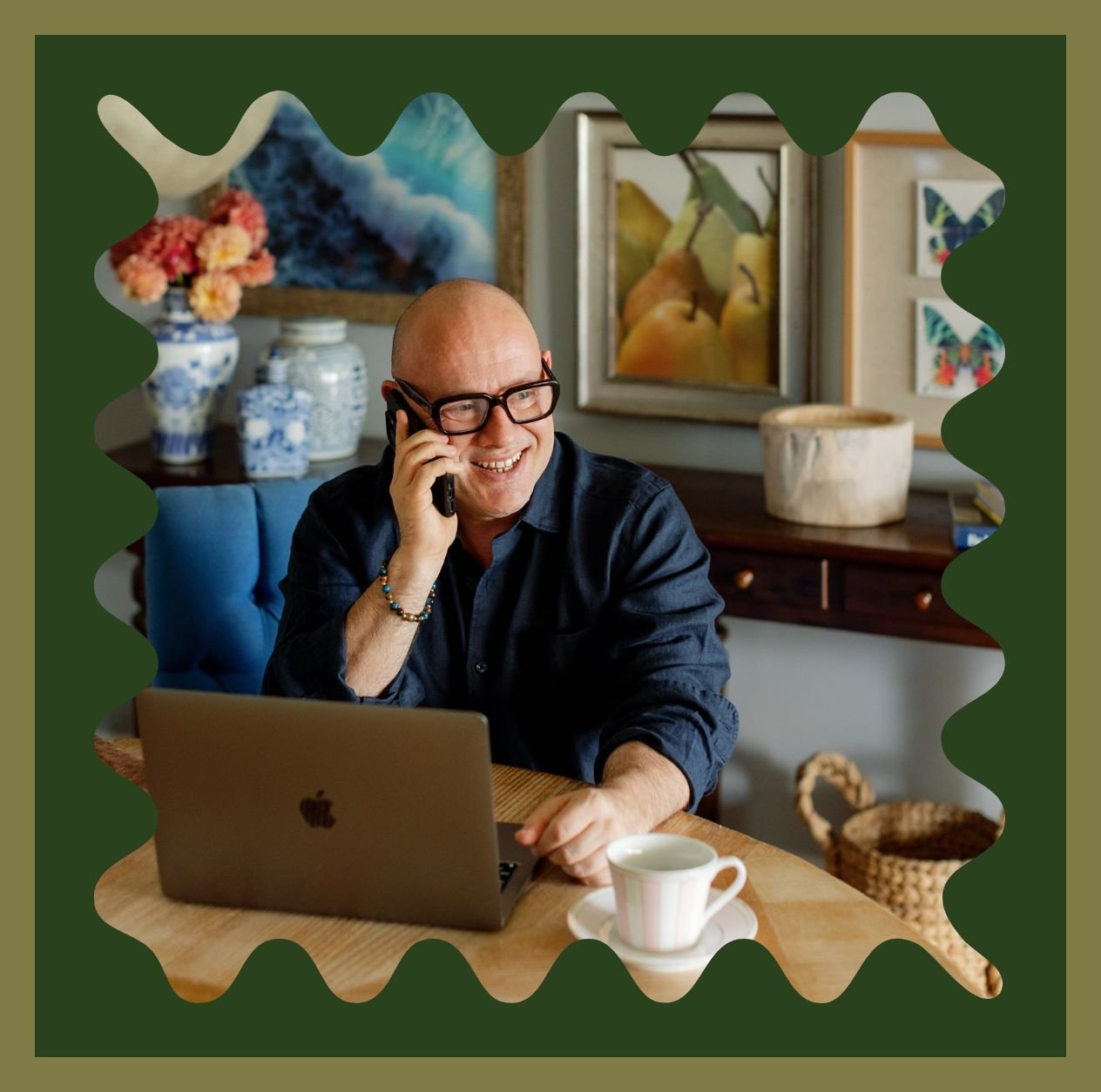 Adam Scougall with glasses talking on the phone at a wooden table with a laptop and a cup of coffee, in a room decorated with paintings, vases, and flowers.