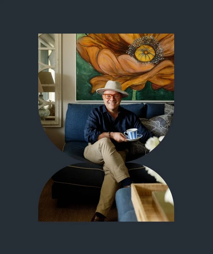 Adam Scougall smiling and sitting on a dark blue couch, wearing glasses, a beige hat, a black shirt, and beige pants. They are holding a small blue and white cup. Behind them is a large colorful painting of a flower.
