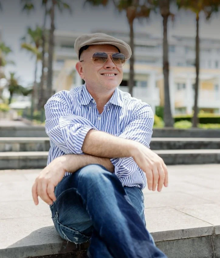 Adam Scougall outdoors on concrete steps, smiling, wearing sunglasses, a beige cap, a blue and white striped shirt, and jeans, with palm trees and buildings in the background.