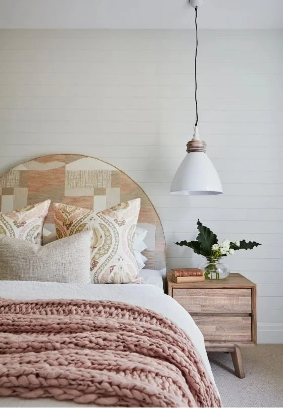 A bedroom scene with a bed, patterned pillows, a pink chunky knit blanket, a wooden nightstand with a glass vase of flowers, and a hanging white pendant light.