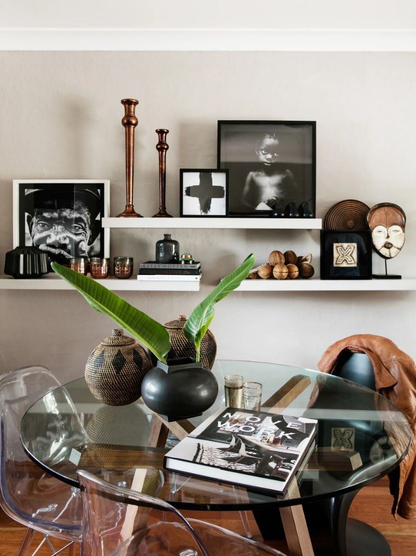 Modern living room with a glass dining table, decorative vases, and framed black-and-white art on wall shelves.