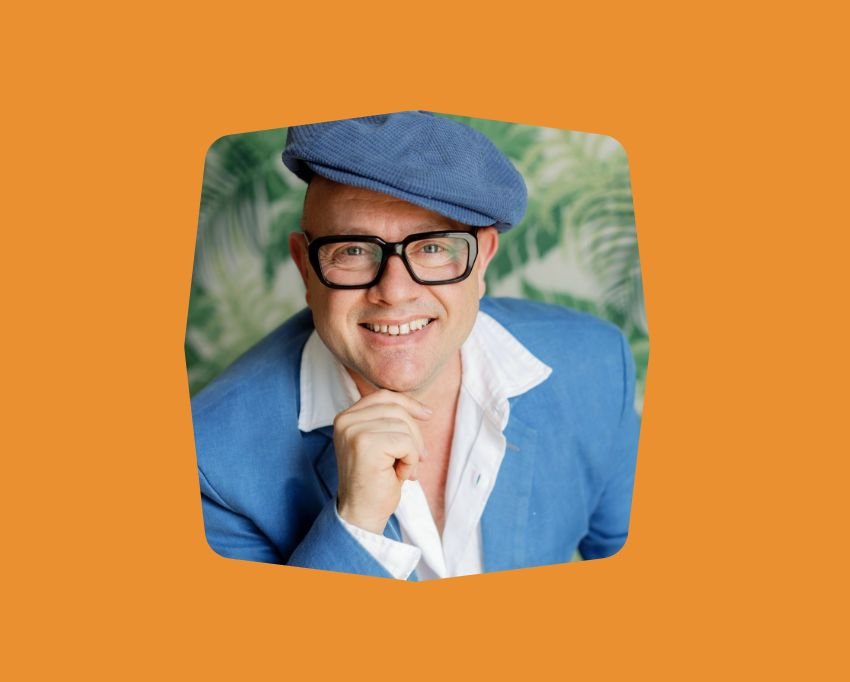 Portrait of a Adam Scougal wearing glasses, a blue beret, a white shirt, and a blue blazer, with a green leafy background.