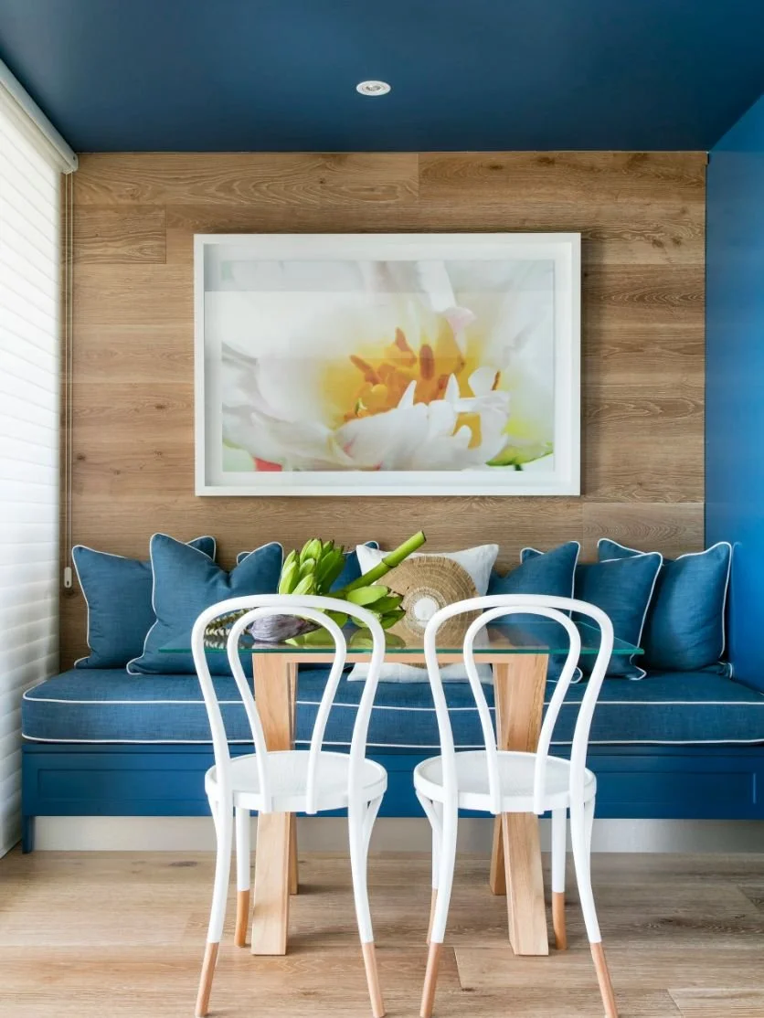 Interior of a cozy room with a blue cushioned bench, navy pillows, a small wooden table, white chairs, a glass vase with tulips, and a framed flower photograph on a wooden wall.