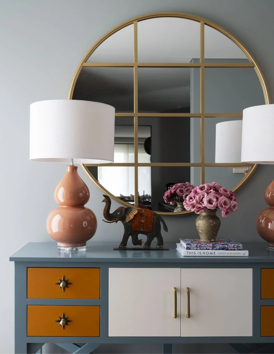 Decorative sideboard with a large round mirror, two pink table lamps, a small elephant figurine, a vase of pink flowers, and books.