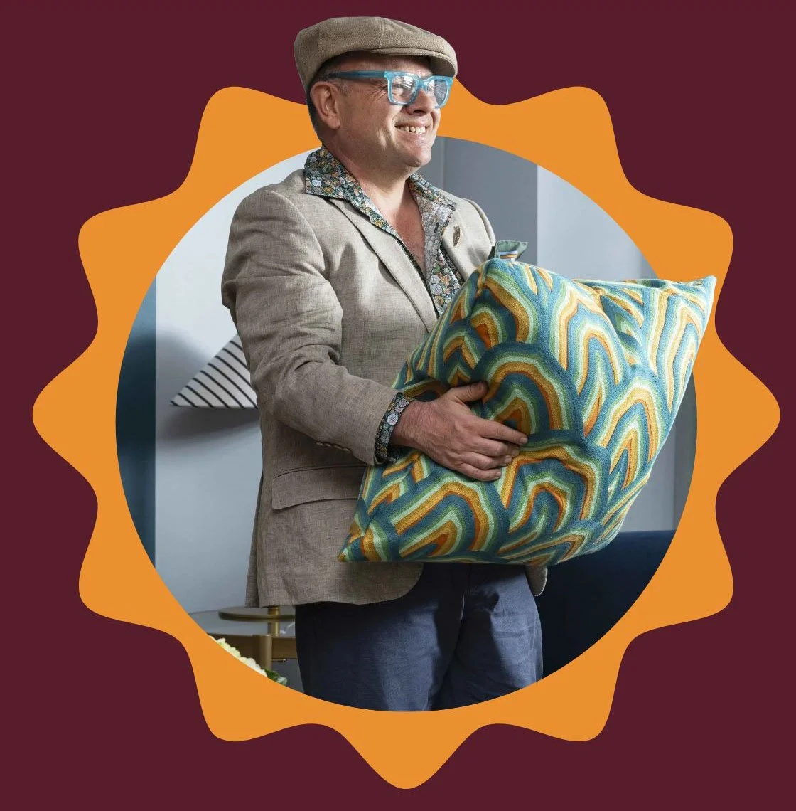 Adam Scougall wearing glasses, a beige hat, a beige blazer, and a colorful shirt holds a large, brightly patterned pillow.