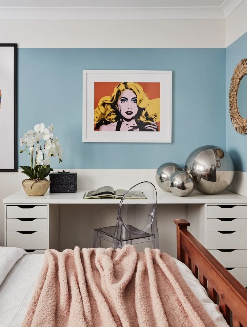 Decorative bedroom corner with a white desk, a transparent chair, a pink blanket on the bed, a white orchid, metallic spheres, and colorful artwork on the wall.