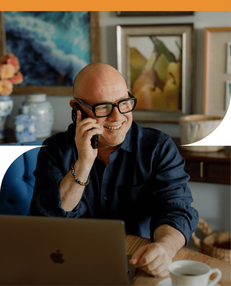 Adam Scougall with glasses and a beaded bracelet, smiling while talking on his cellphone at a desk with a laptop and a cup of coffee in a cozy, decorated room.