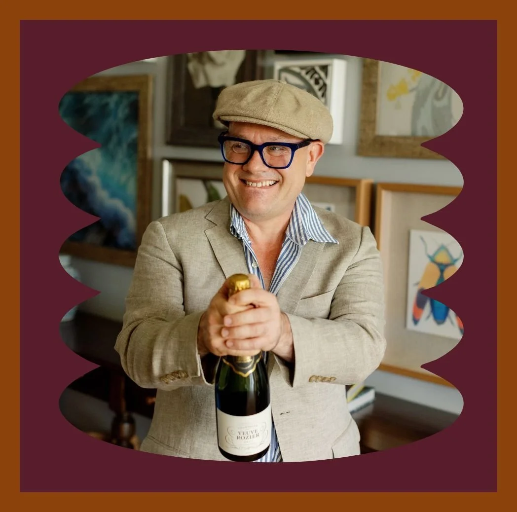 Adam Scougall wearing glasses, a beige flat cap, and a beige blazer, holding a bottle of sparkling wine with a cheerful expression, in an art gallery with paintings on the walls.