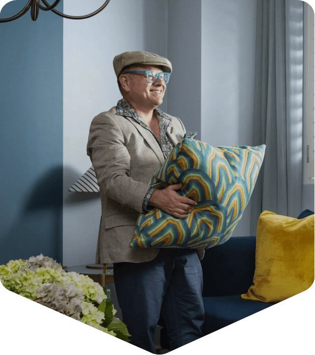 Adam Scougall with glasses, wearing a beige blazer, patterned shirt, and cap, holding a colorful, patterned pillow in a modern living room with blue walls and yellow pillows.