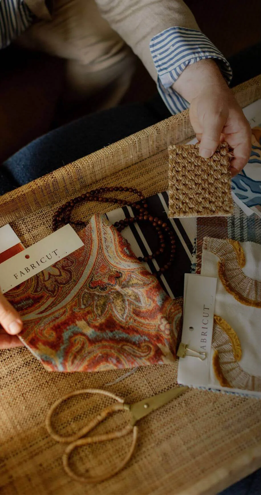 Person holding a textured fabric sample over a table with fabric swatches, a beaded necklace, and a pair of scissors.