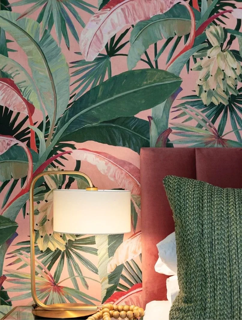 A bedroom corner with a tropical leaf-patterned wallpaper, a pink upholstered headboard, green textured pillow, white bedsheet, a modern table lamp with a gold base, and a beaded necklace on the bedside table.