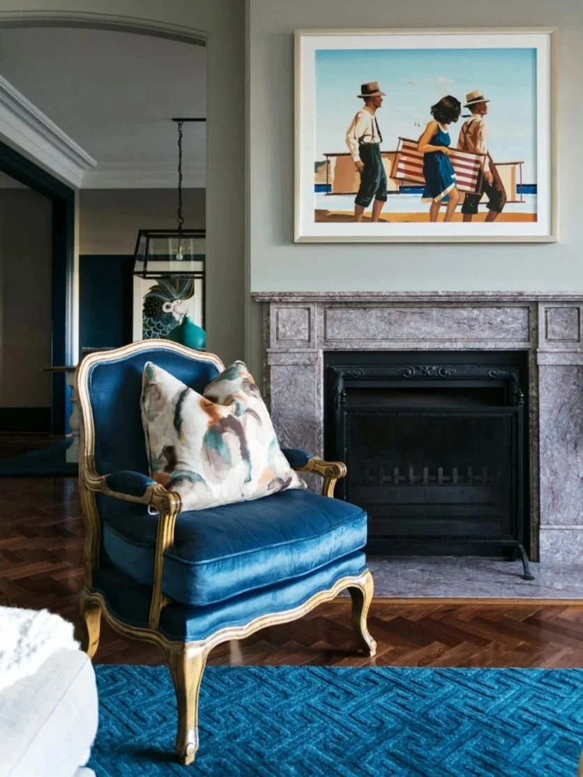 Living room with a blue velvet armchair with a patterned pillow, a fireplace with a marble surround, a blue rug, and a framed painting of children at the beach.