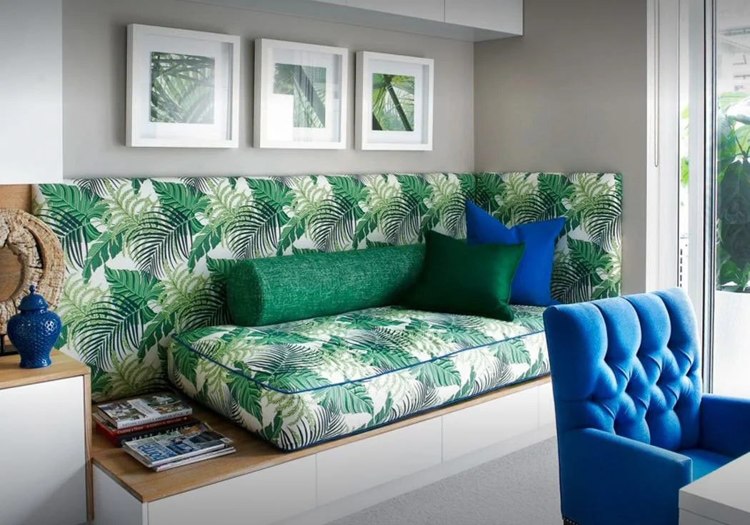 A bright living room with a tropical-themed padded bench and green, blue, and black pillows, a blue tufted chair, a small wooden side table with magazines, and three framed pictures of green palm leaves on the wall next to a sliding glass door.