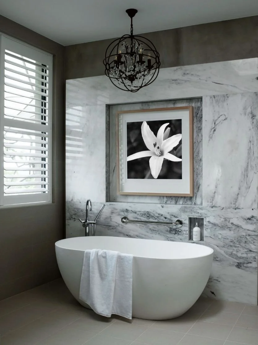 Modern bathroom with a white freestanding bathtub, a black and white flower photograph, marble wall, and a chandelier.