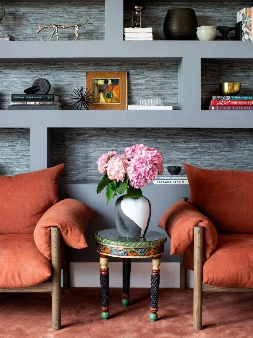 Living room corner with two orange armchairs, a small colorful side table, and a vase of pink hydrangeas on top. Shelves with decorative items and books are in the background.