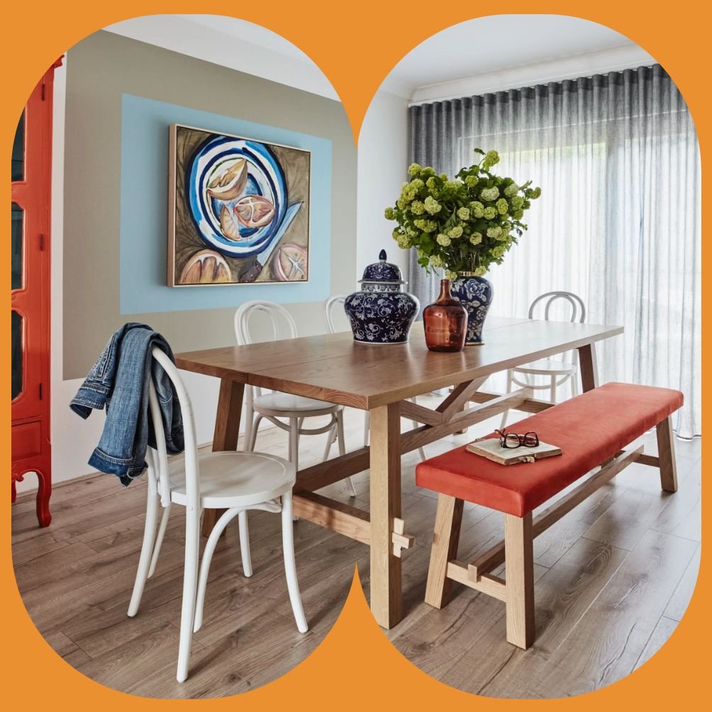 A dining room with a wooden table, white chairs, and a red bench. Decor includes vases with flowers, a book, and eyeglasses on the bench. Artwork hangs on the wall, and a window with gray curtains allows natural light into the room.