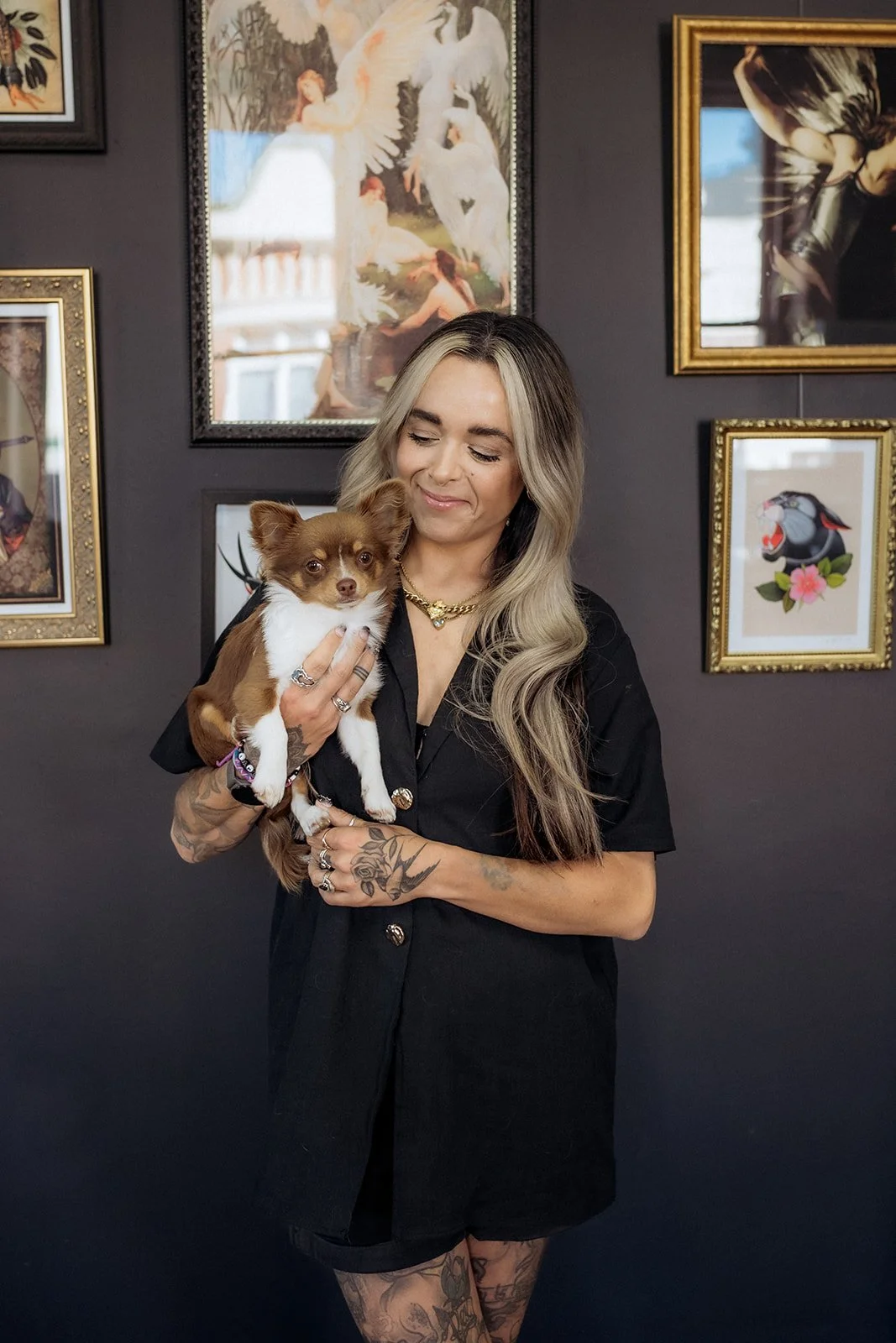 Smiling tattooed woman in a black shirt gently holding a small brown and white dog against a gallery wall of framed artwork