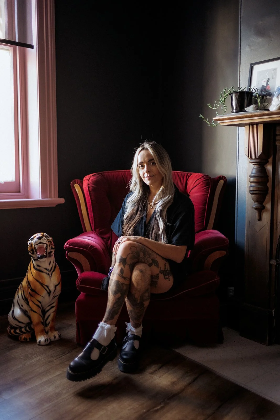 Tattooed woman seated on a red armchair in studio beside a decorative tiger statue and fireplace