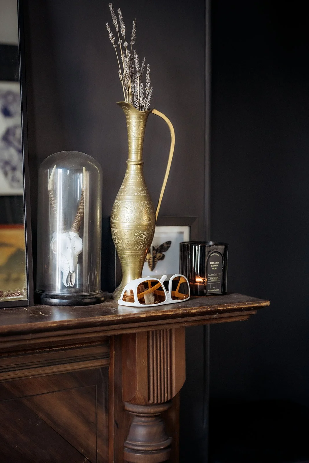 Decorative studio shelf with a gold vase of dried flowers, a glass dome displaying a skull, protective eyewear, and a lit candle against a dark wall