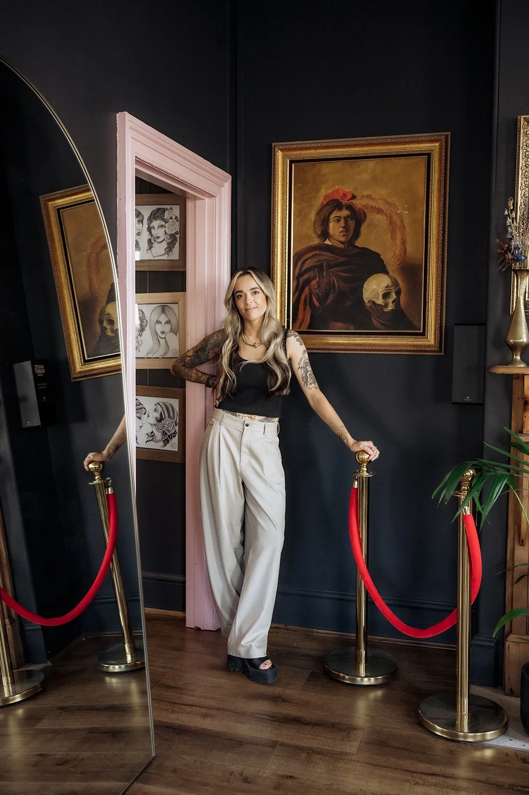 Tattooed woman in a black top and beige trousers leaning against a velvet rope barrier beside framed artwork and a large mirror in a moody studio space
