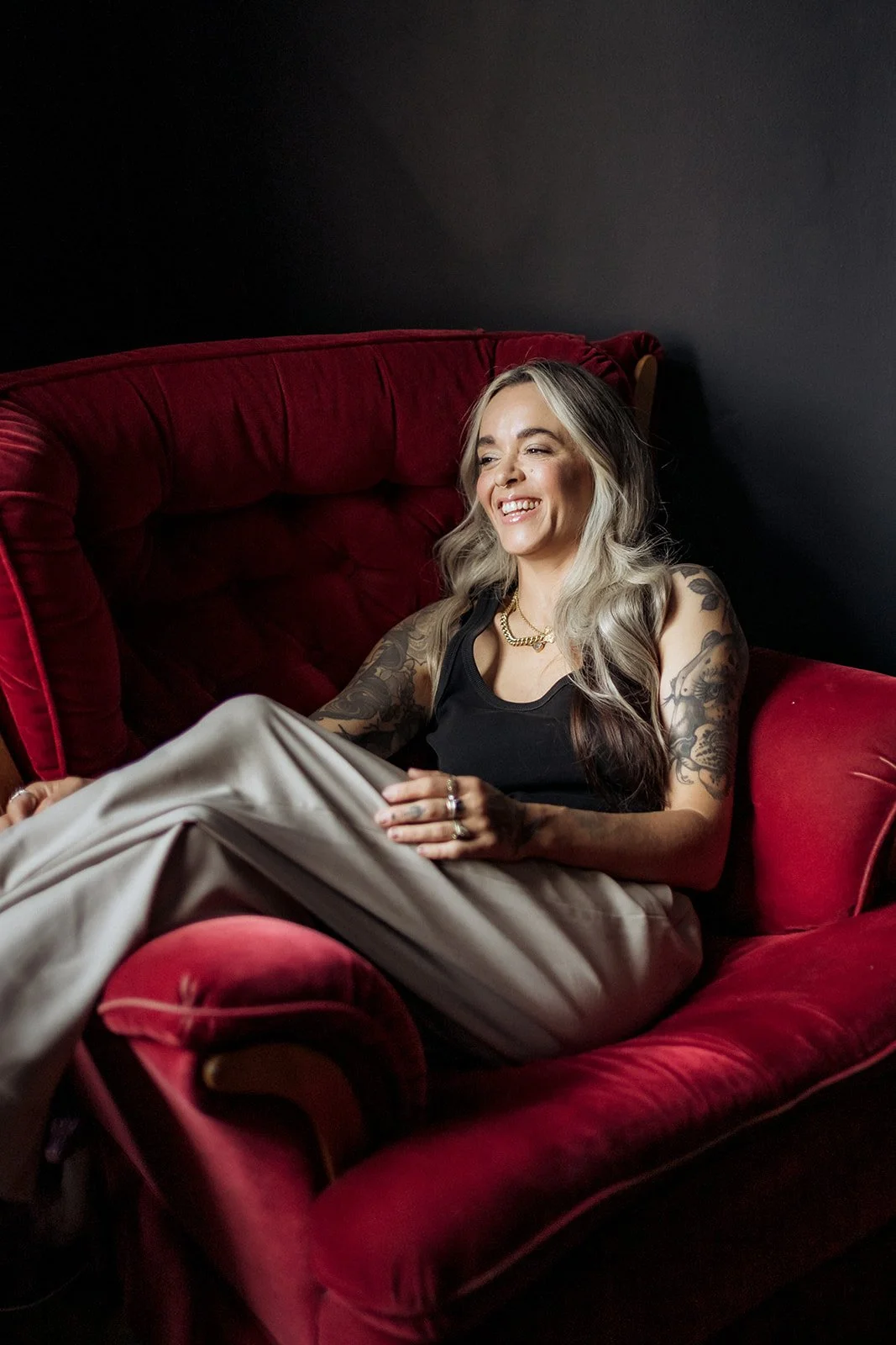 Woman with tattooed arms sitting relaxed in a deep red velvet armchair, smiling in a softly lit studio setting
