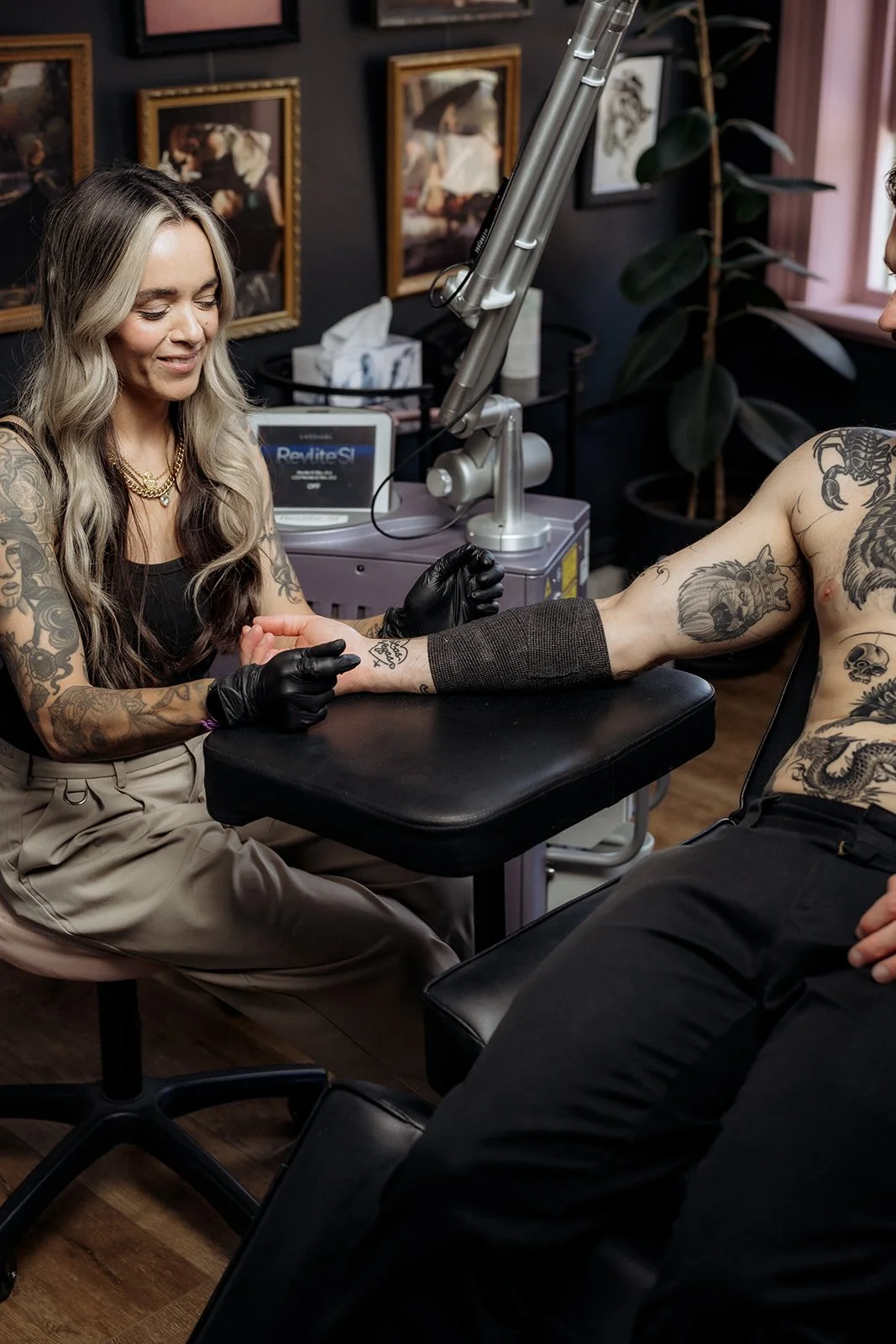 Tattoo removal practitioner in gloves examining a client’s tattooed forearm beside a laser machine in a studio with framed artwork