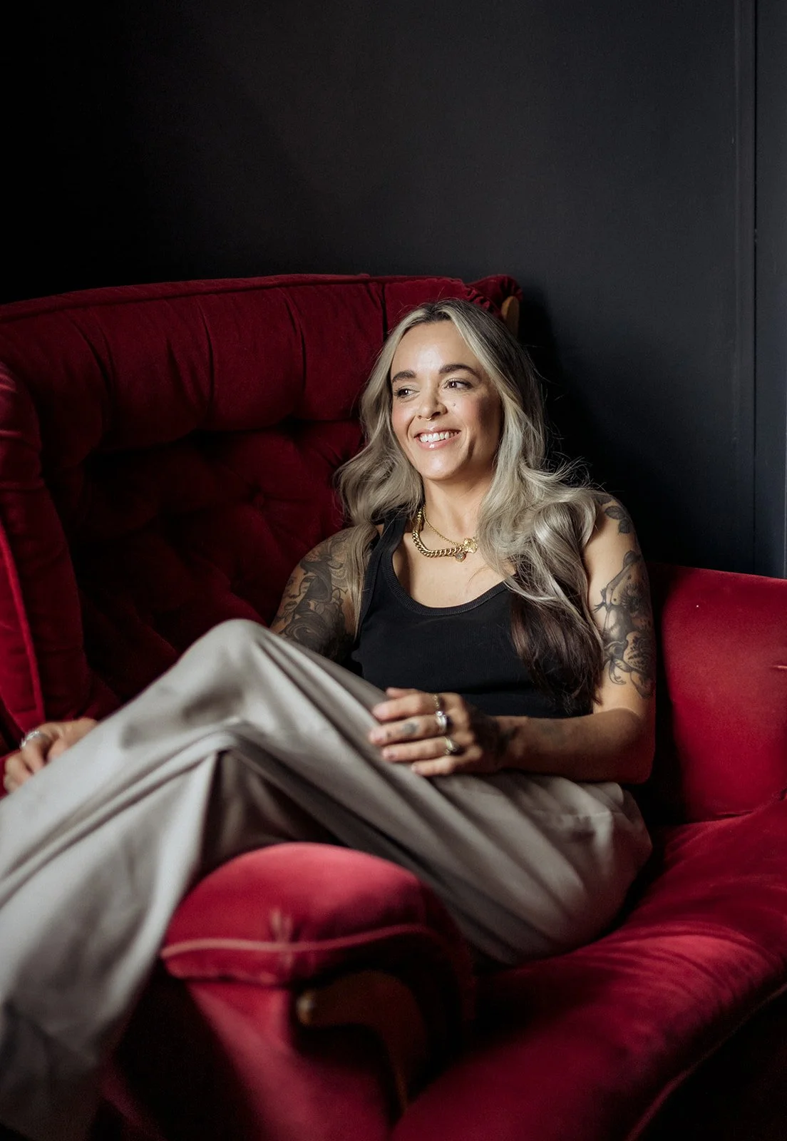 Woman with tattooed arms sitting relaxed in a deep red velvet armchair, smiling in a softly lit studio setting