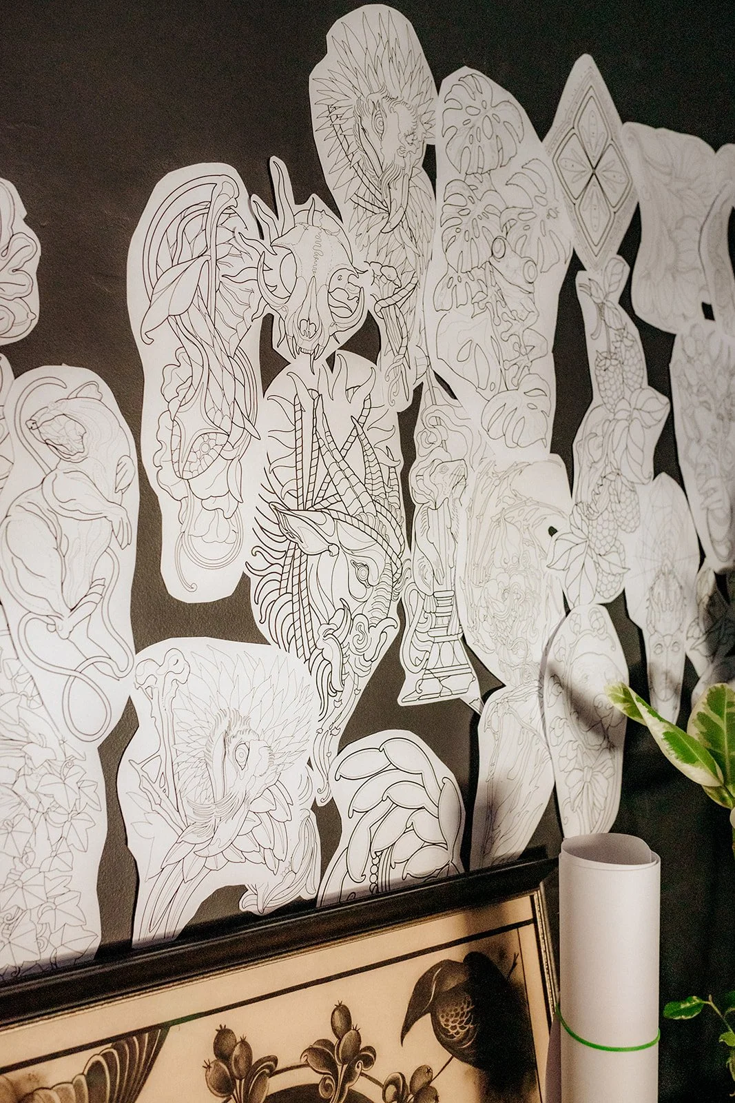 Selection of detailed tattoo flash designs displayed on a dark studio wall, with framed artwork and greenery below