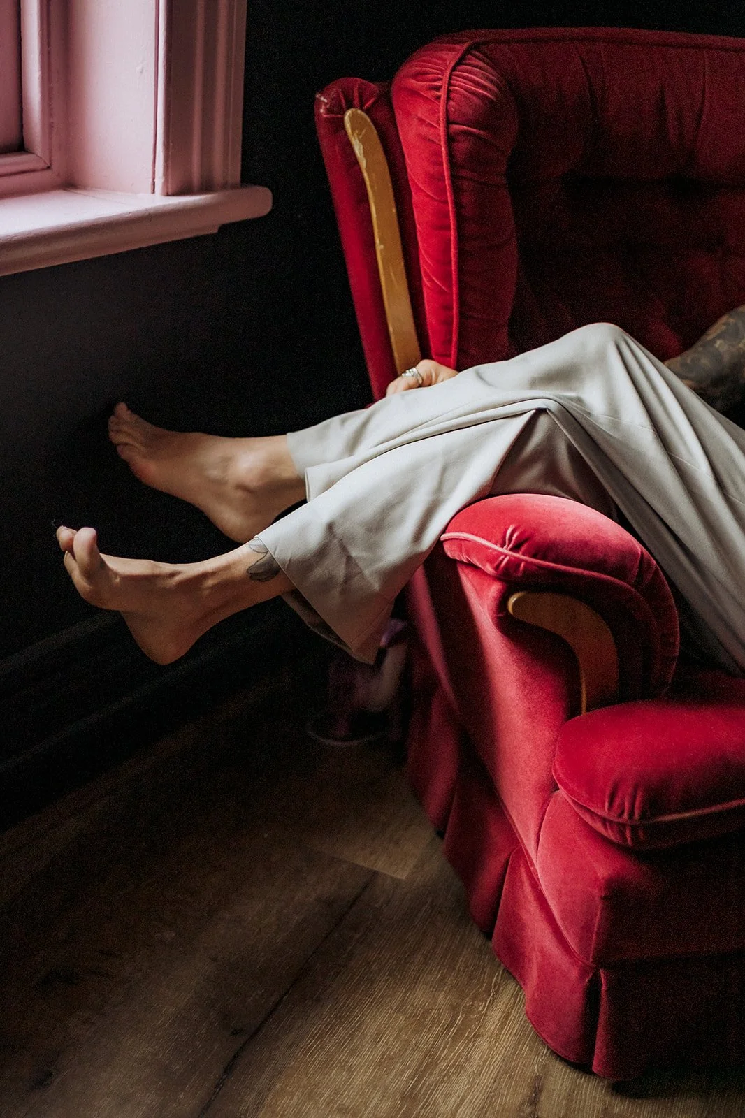 Bare feet resting on the arm of a plush red velvet chair beside a window, with soft natural light and wooden flooring