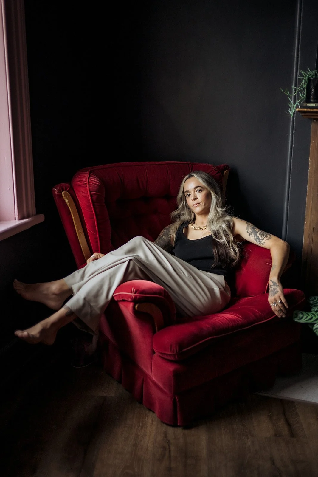 Tattooed woman reclining barefoot on a deep red armchair in a softly lit studio, wearing a black top and beige trousers