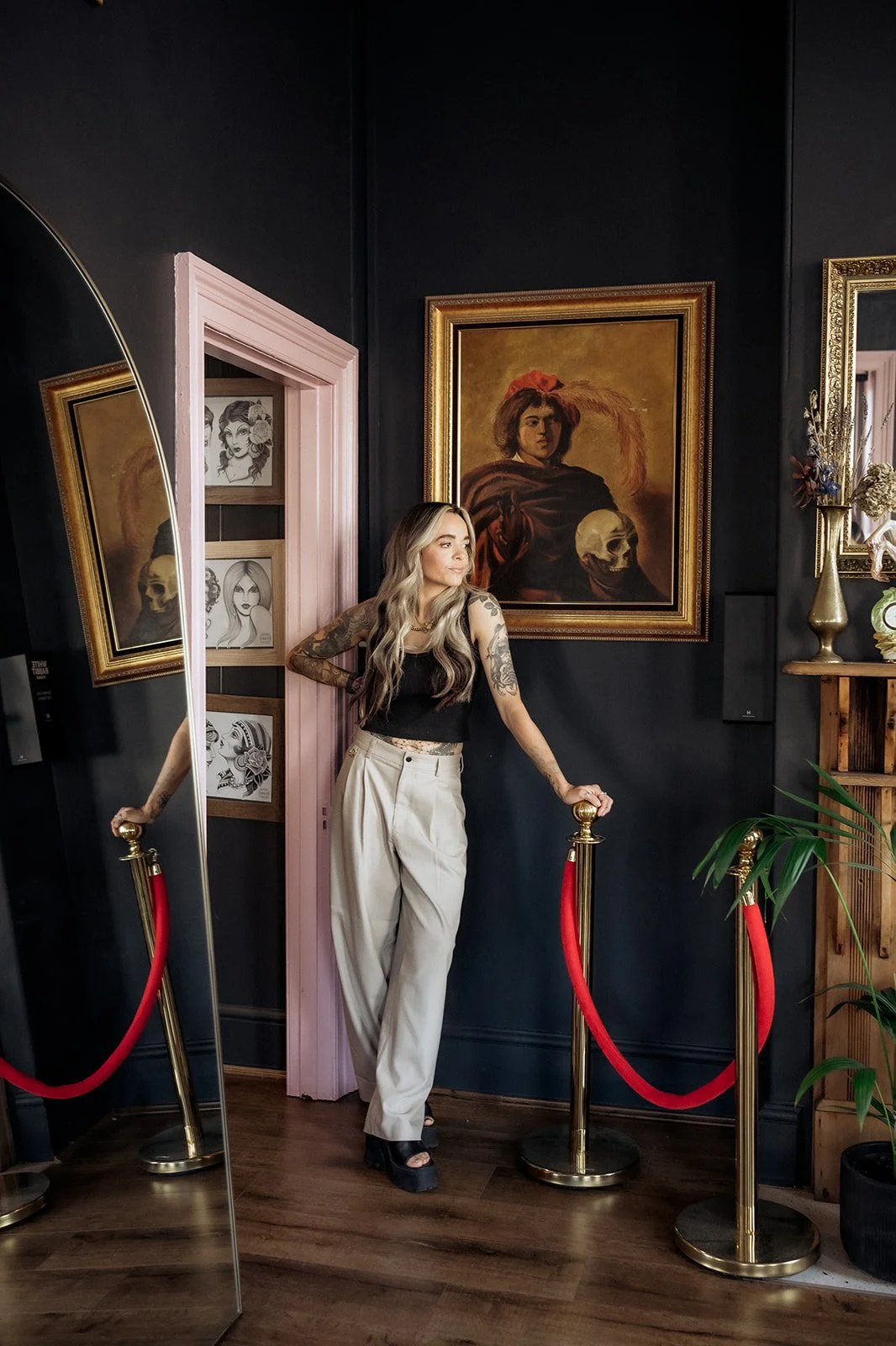 Tattooed woman in a black top and beige trousers leaning against a velvet rope barrier beside framed artwork and a large mirror in a moody studio space