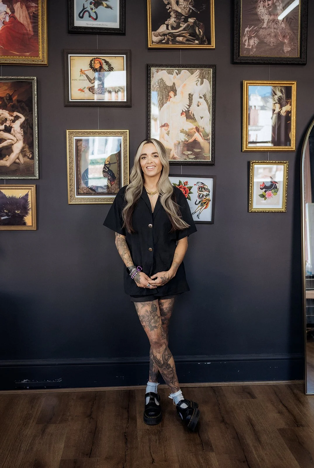 Smiling tattooed woman standing in studio against a gallery wall of framed artwork